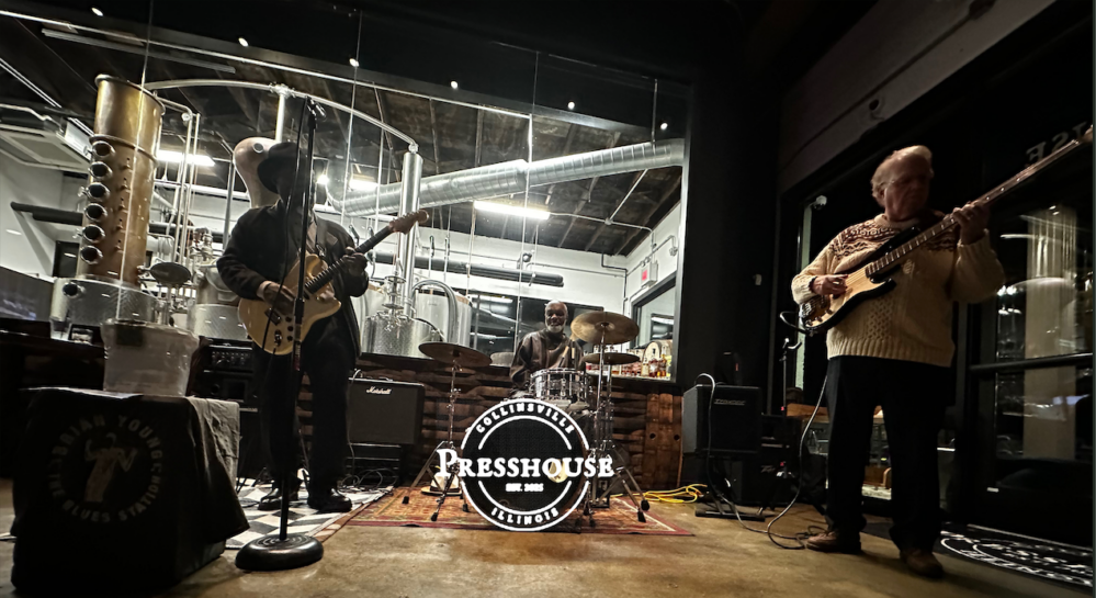 Two men playing guitars and one man playing drumset in front of distilling equipment at PressHouse in Uptown Collinsville