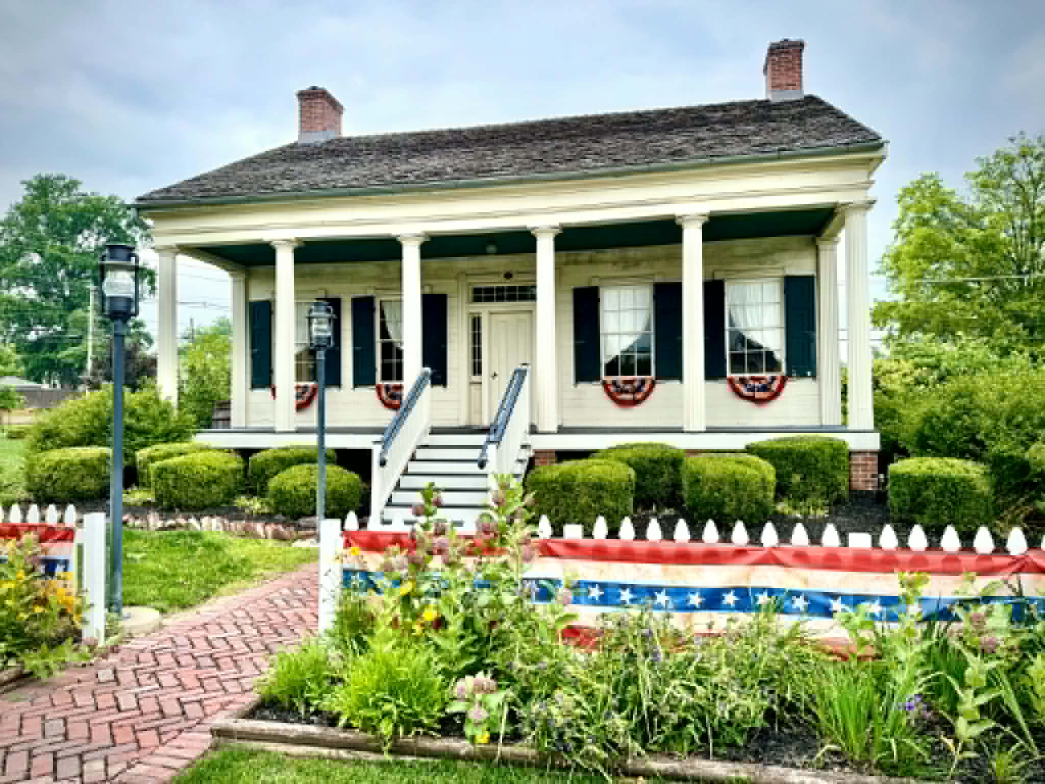 Image of historic DD Collins House, a small white house with American bunting and decorations and a brick paver sidewalk.